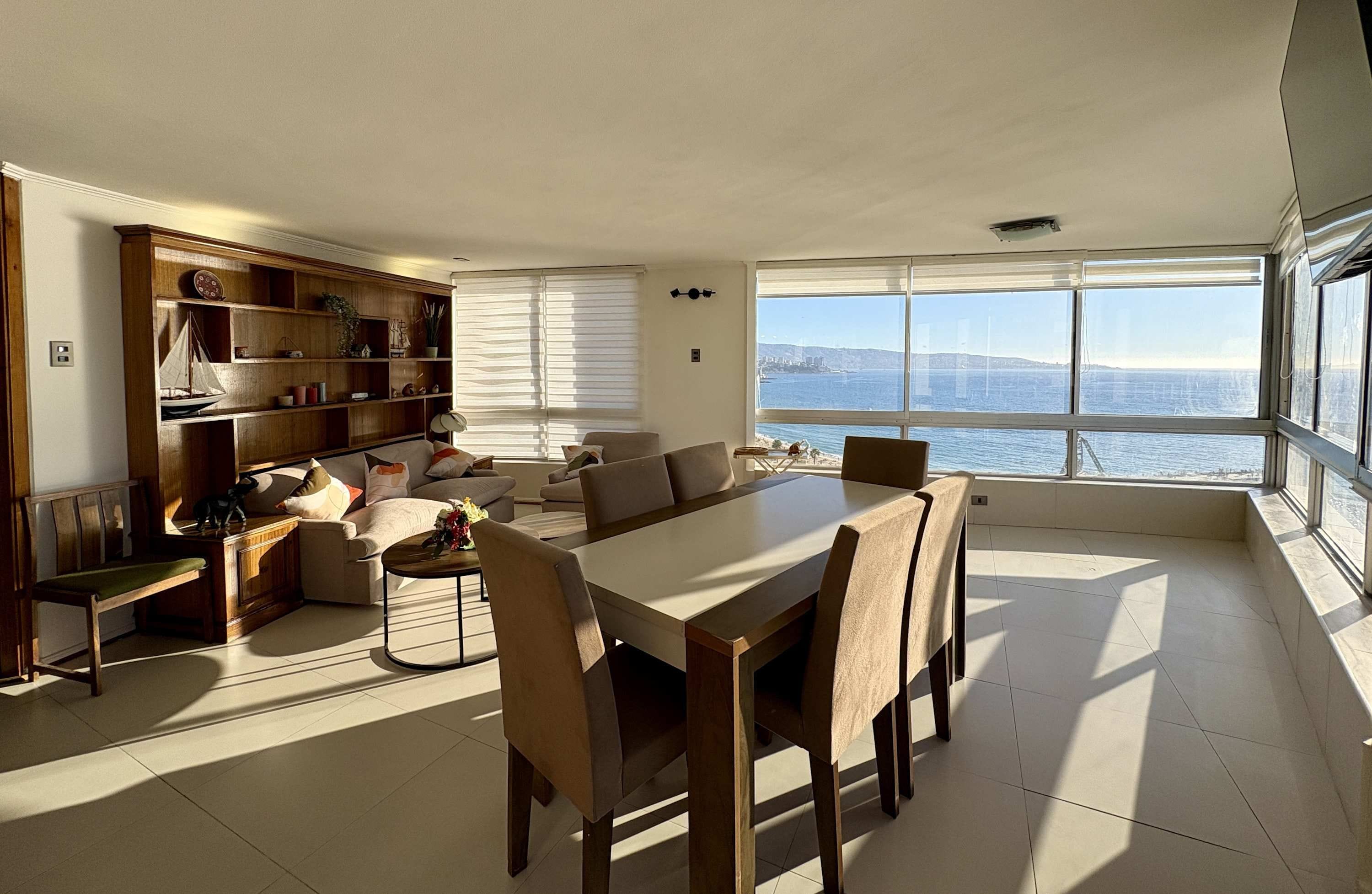 Amplia sala de estar con una gran mesa de comedor, asientos cómodos, una estantería de madera y vistas al mar a través de amplios ventanales.