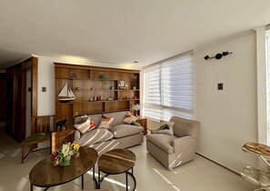 Ein Wohnzimmer mit einer Couch, zwei Couchtischen, einem Bücherregal und einem Fenster mit Jalousien.