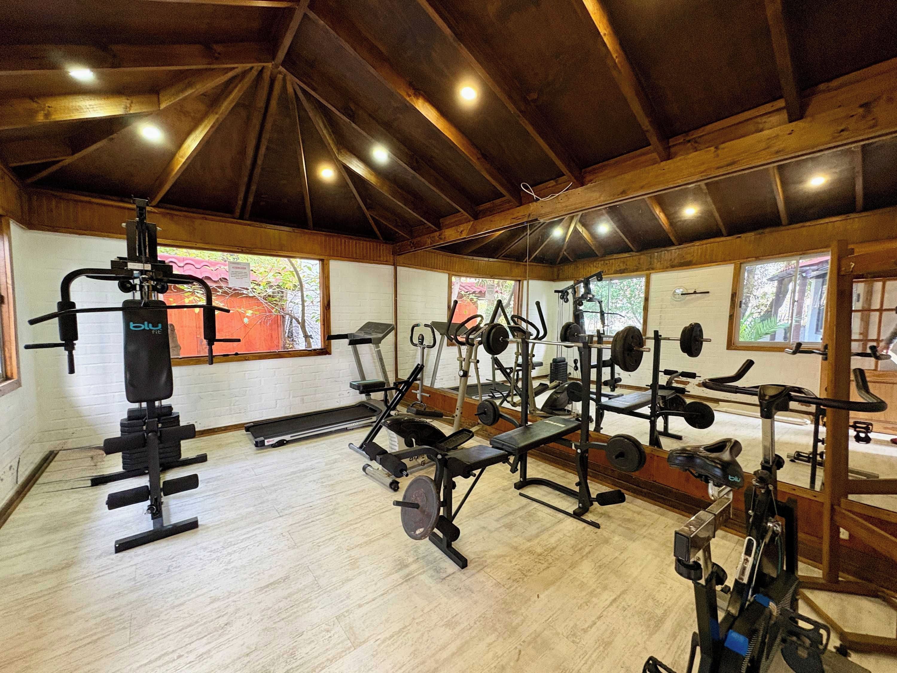 Un gimnasio con diversos equipos de ejercicio, vigas de madera en el techo y ventanas amplias.