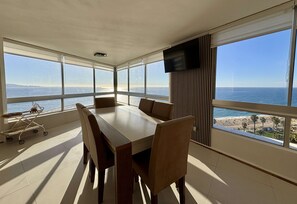 Quarto com uma mesa grande e cadeiras, uma TV na parede e vista para o mar.