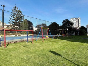 Ein von einer Zäunung umgebener Spielplatz mit Schaukeln und einem Fußballtor, umgeben von Gras und Bäumen, mit Gebäuden im Hintergrund.