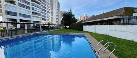 Una piscina con una escalera de metal, rodeada por un muro blanco y una zona cubierta de césped.
