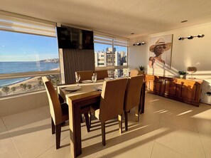 Uma sala de jantar com vista para a praia e para a paisagem urbana, uma mesa de jantar de madeira e um espaço de bar com uma planta e objetos decorativos.