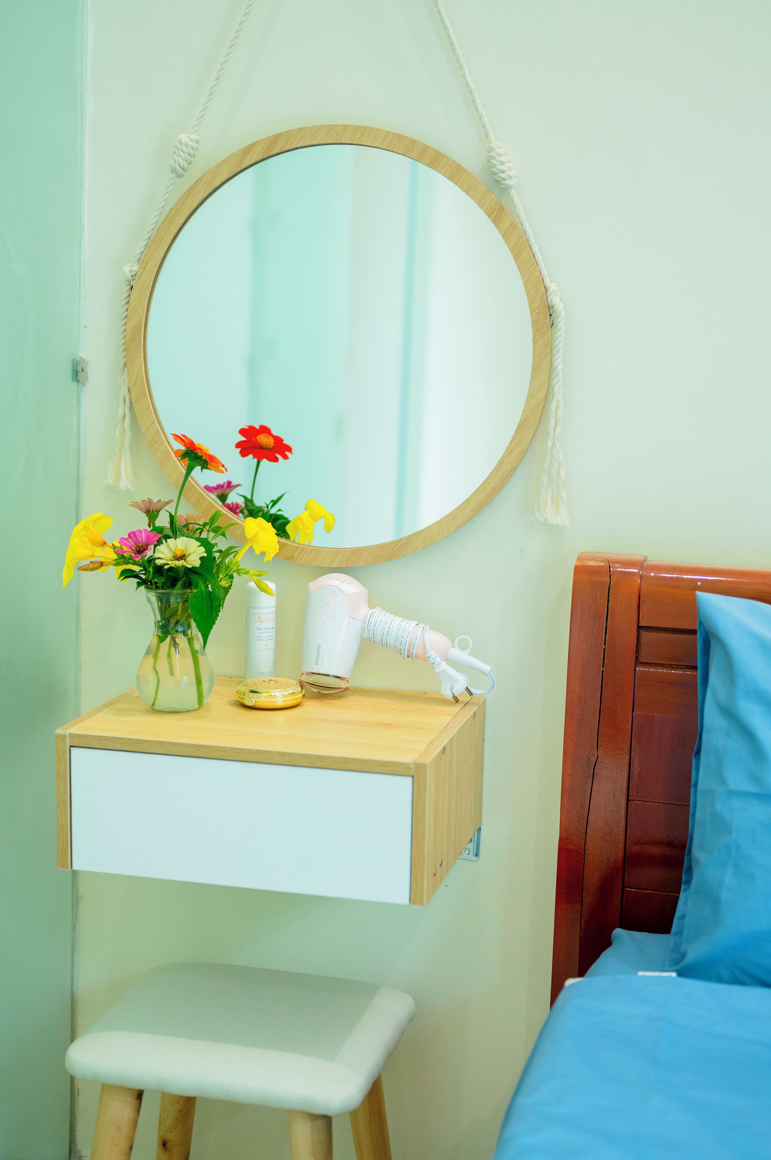 Quarto com mesa de cabeceira de madeira, espelho, um pequeno banquinho, cama com cobertura azul e prateleira na parede com artigos de higiene pessoal.