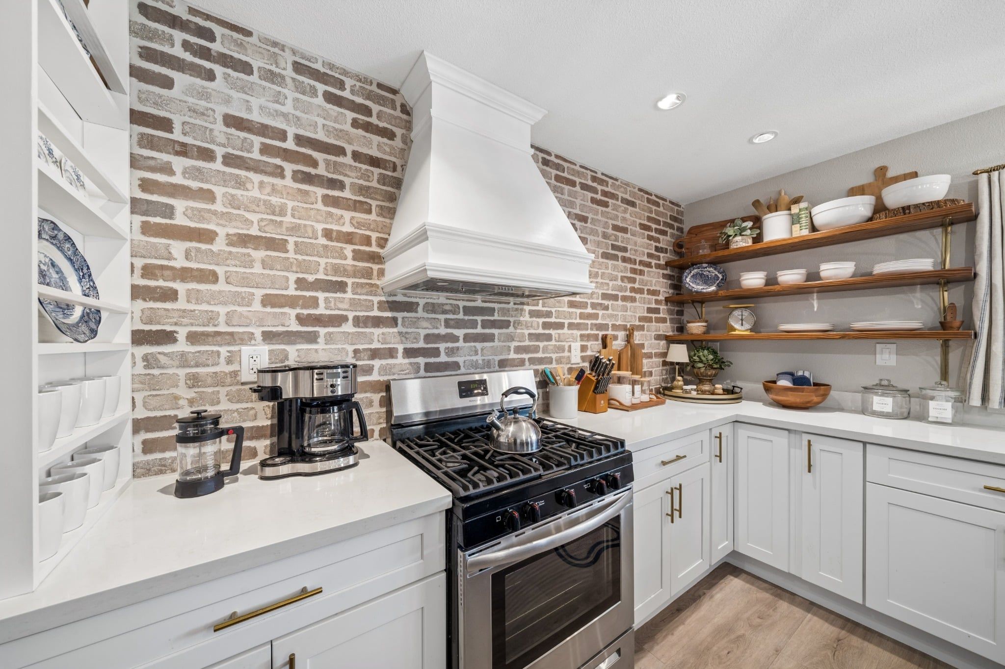 A kitchen with a brick wall, white cabinets, a gas stove, and various kitchen utensils.
