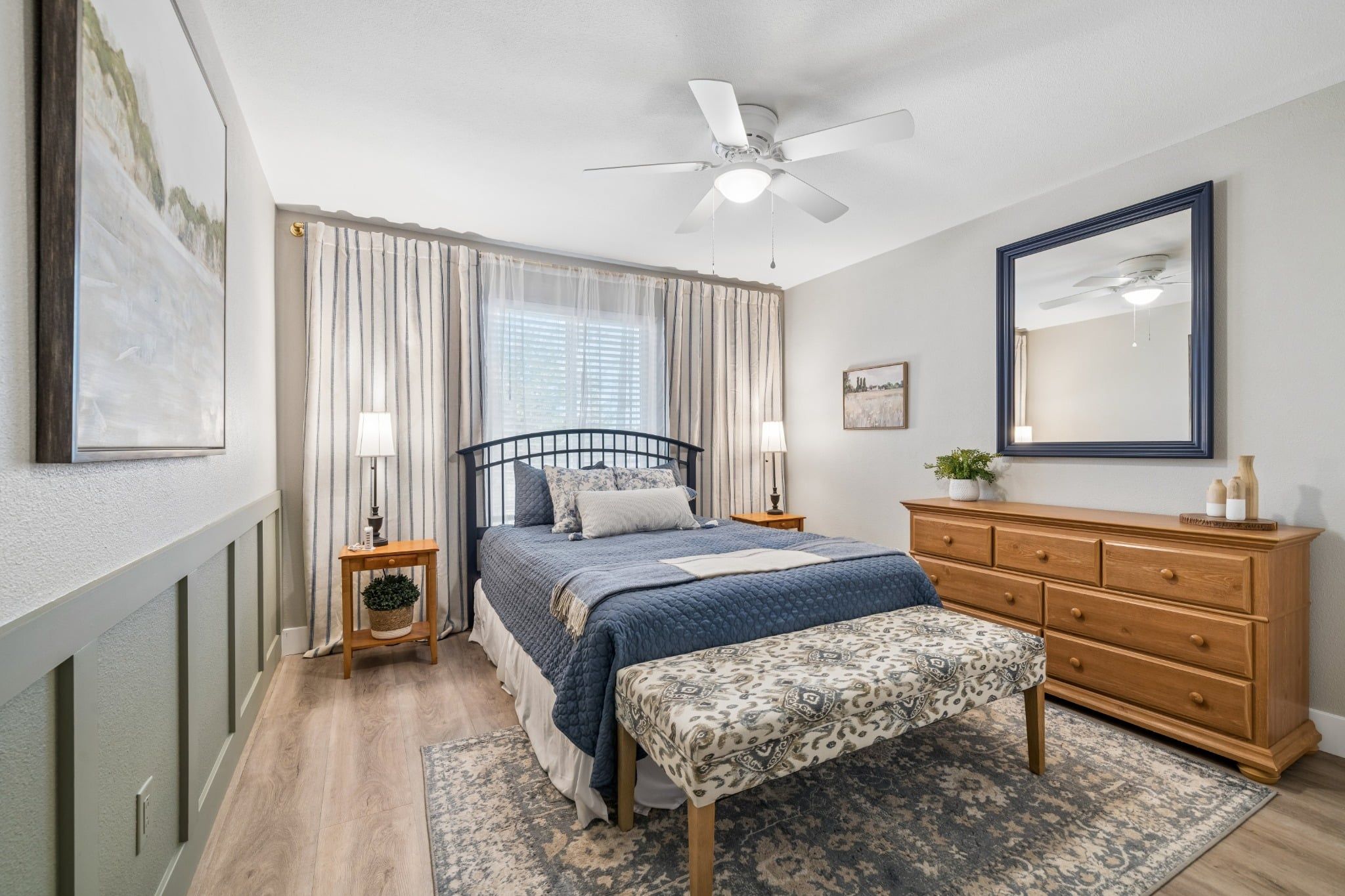 A bedroom with a bed, bedside tables, a dresser, a ceiling fan, and a window with curtains.