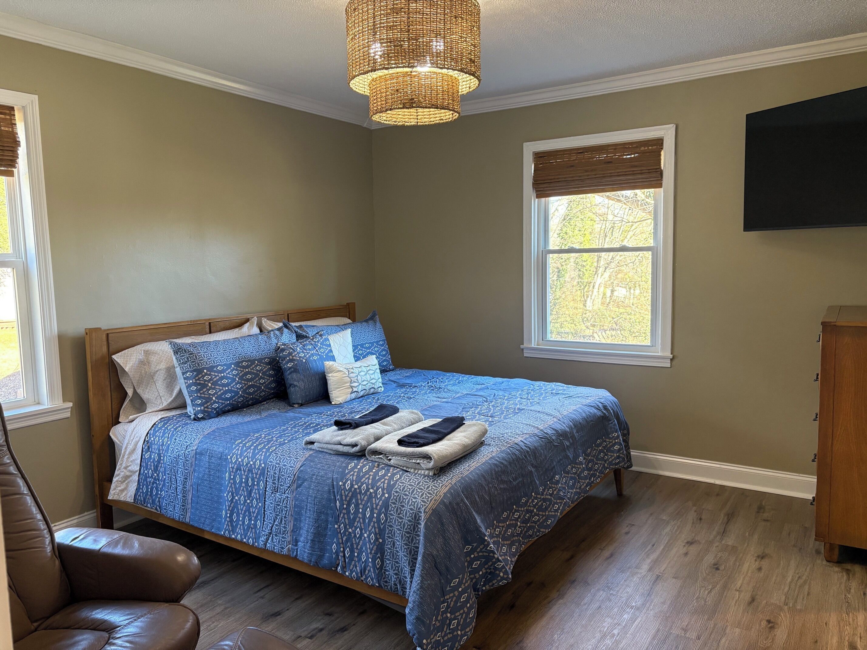 A bedroom with a bed, a wooden headboard, a window with blinds, a brown leather chair, and a television mounted on the wall.