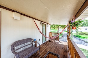 Eine hölzerne Veranda mit einer Korbmöbelbank und einem Schaukelstuhl, einer roten und weißen Seilsperre sowie einem hängenden Blumentopf.