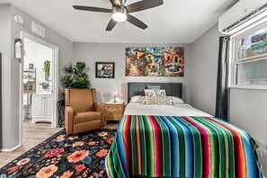 A bedroom with a bed, a chair, a nightstand, a ceiling fan, a window, and a painting on the wall.