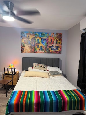 A bedroom with a bed, a nightstand, a chair, a ceiling fan, and a painting on the wall.