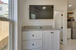 A kitchen with white cabinets, a granite countertop, a flat-screen TV mounted above the countertop, and a stainless steel refrigerator. - #7 | Modern SPI Beach Studio + Pool | Smart TV (South Padre Island)