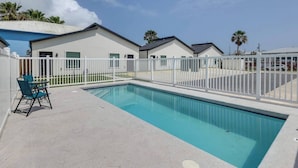 A swimming pool with a white fence, a table with chairs, and buildings in the background. - #7 | Modern SPI Beach Studio + Pool | Smart TV (South Padre Island)