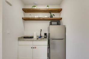 A compact kitchen with a stainless steel refrigerator, a microwave, and a coffee maker on the counter. There are two white cabinets and a wooden shelf above with potted plants. - #7 | Modern SPI Beach Studio + Pool | Smart TV (South Padre Island)