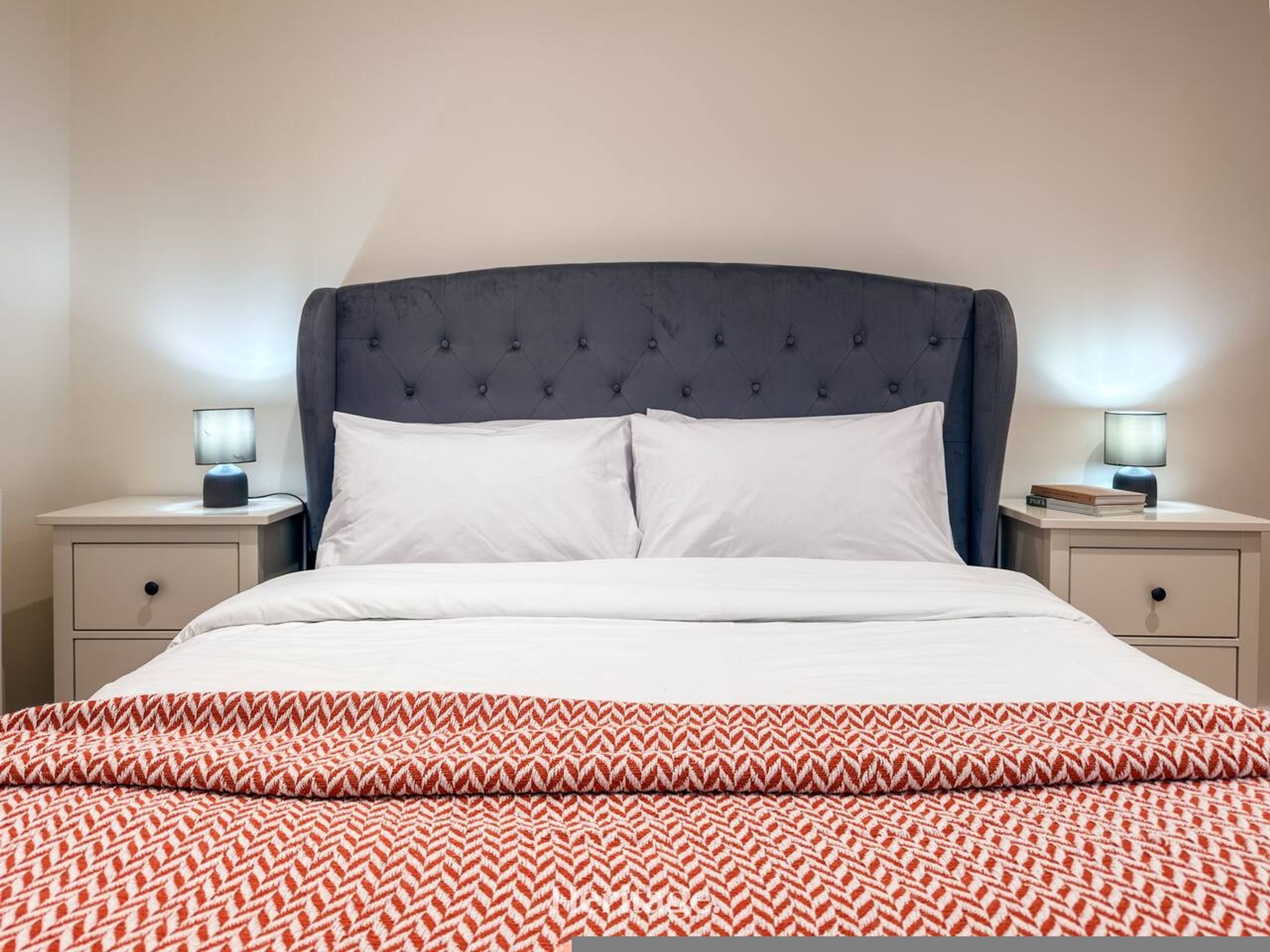 Una cama con cabecera capitonada, dos mesitas de noche con lámparas y una colcha estampada.