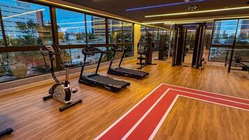 Ein Fitnessstudio mit Laufbändern, Trainingsgeräten und großen Fenstern mit Blick auf die Stadt.