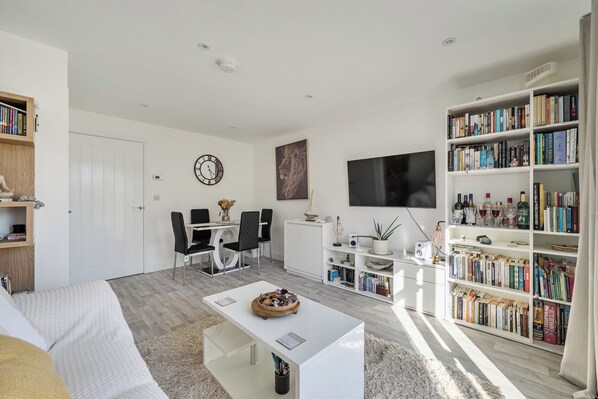 A modern living room with a dining area, a bookshelf filled with books, a flat-screen TV, and a decorative clock.