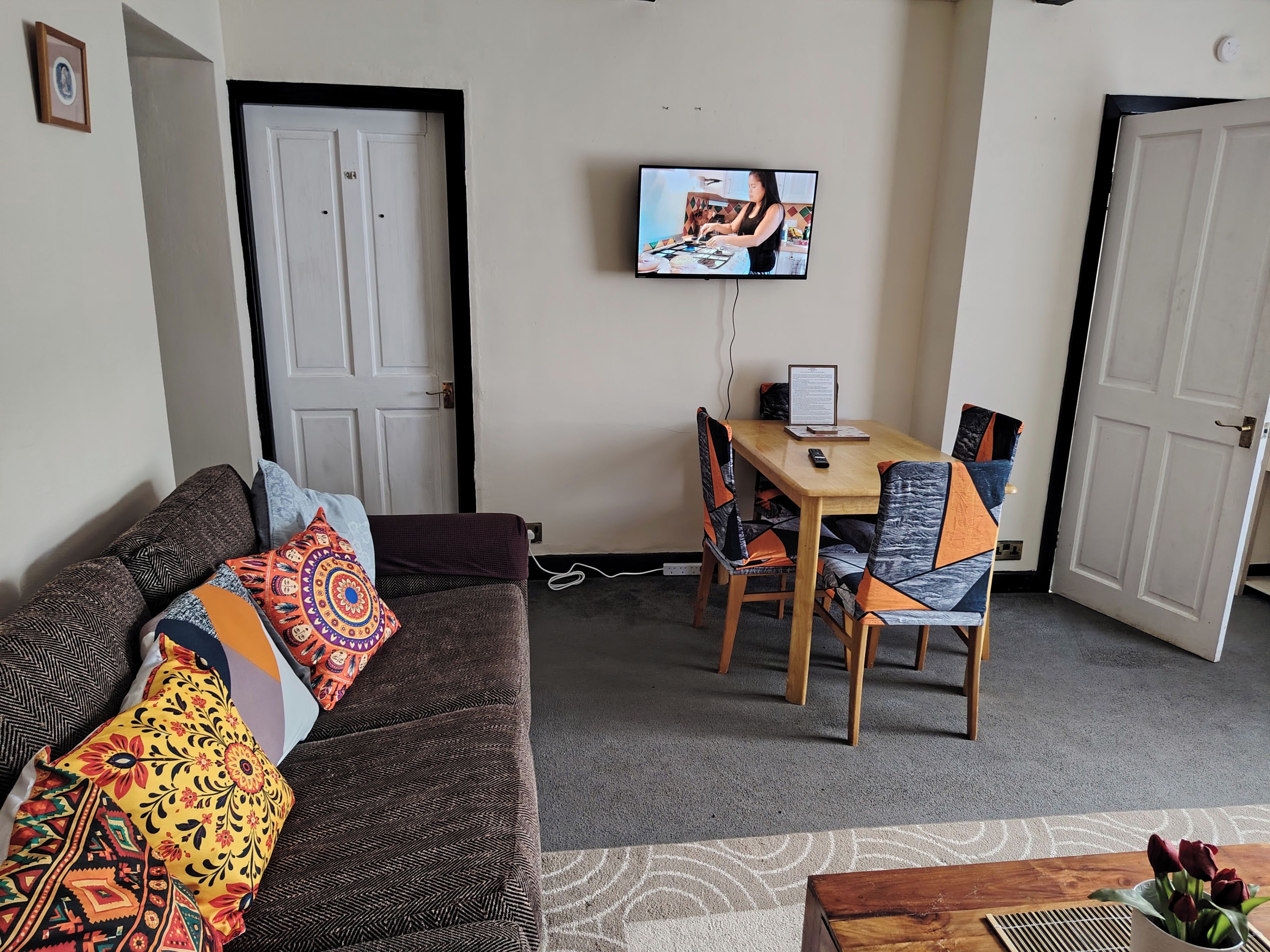 Una sala de estar con un sofá, una mesa de comedor con sillas, un televisor montado en la pared y una puerta.