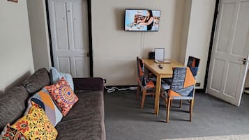 Una sala de estar con un sofá, una mesa de comedor con sillas, un televisor montado en la pared y una puerta.