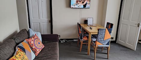 Una sala de estar con un sofá, una mesa de comedor con sillas, un televisor montado en la pared y una puerta.