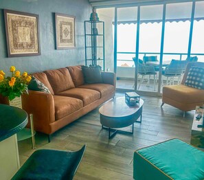 A living room with a brown sofa, a round coffee table, and a teal accent chair. There are framed artworks on the wall and a view of the outdoors through large windows. - Boana Torre Malibu #803 , Ocean view, romantic zone (Puerto Vallarta)