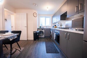 A compact kitchen with a washing machine, a dining area with a table and chairs, and a clock on the wall. - Cozy Coventry Retreat - 3 Bedroom House (Coventry)