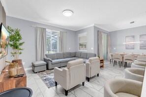 A modern living room with a sectional sofa, a dining area with chairs, and a large window with curtains. - Renovated 4 BDR With pvt Pool (Kissimmee)