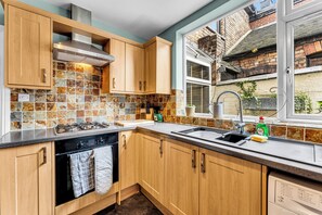 A kitchen with wooden cabinets, a tiled backsplash, a stainless steel range hood, and a window with a view of a brick building. - Spacious Stoke Townhouse 3Br Near Hospital (Stoke-on-Trent)