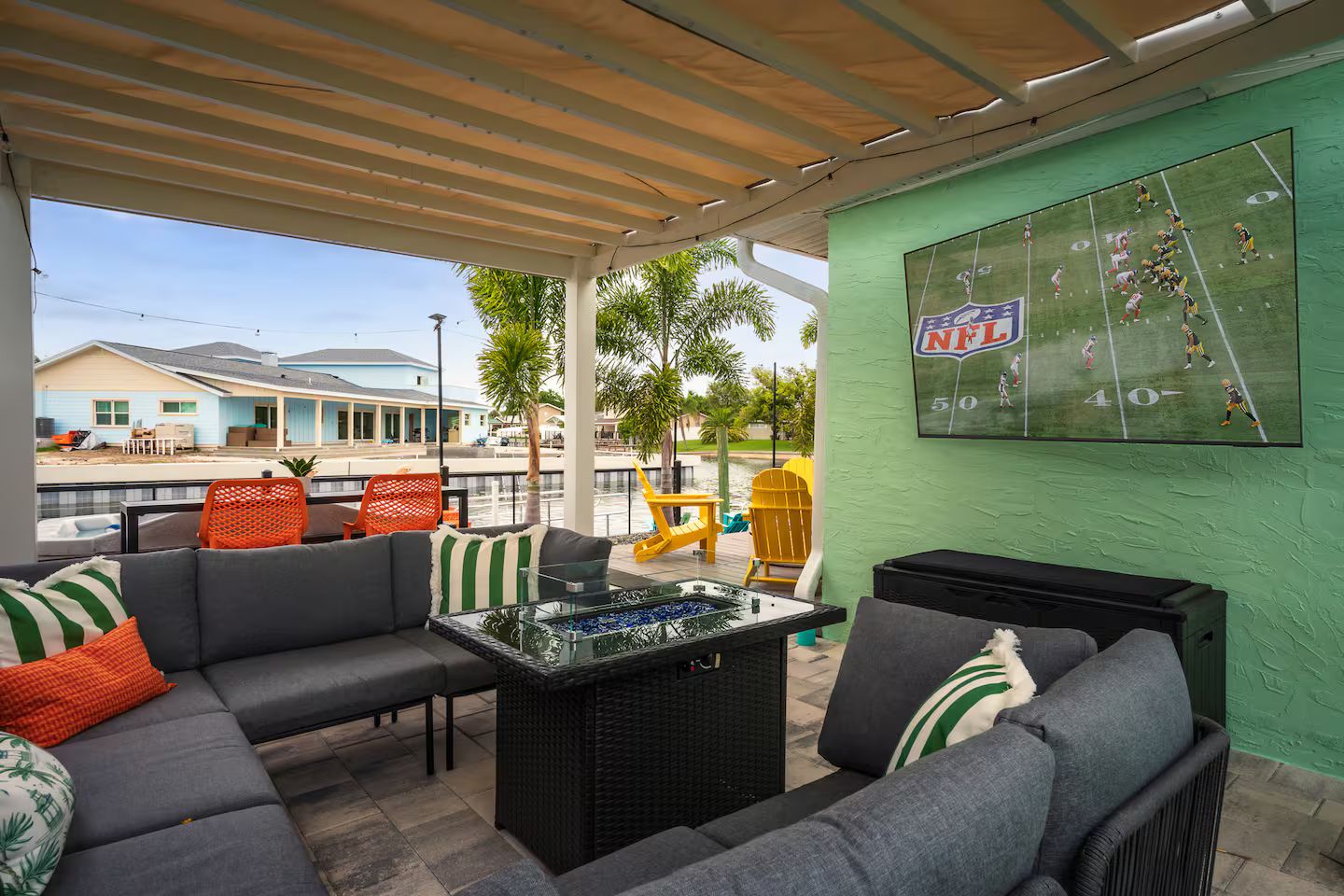 A covered outdoor seating area with a pool table, sofas, and a large screen displaying a football game.