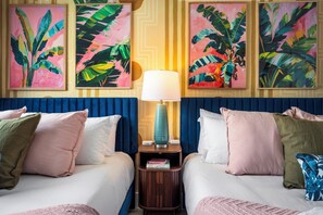 Ein Hotelzimmer mit zwei Betten, einem Nachttisch mit Lampe und Wandbildern mit tropischem Motiv.