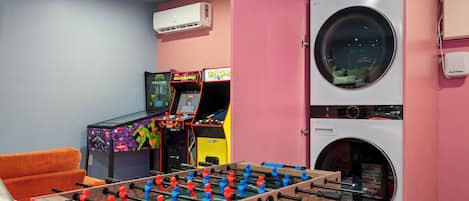 Una habitación con mesa de futbolín, juegos de arcade y lavadora.