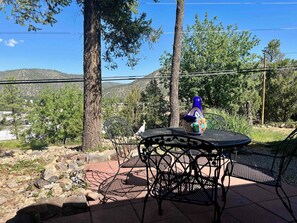 An outdoor patio with a table and chairs, surrounded by trees and a view of a hilly landscape. - Groovy View Cabin- Mountain Views W Covered Porch - Pet Friendly (Ruidoso)