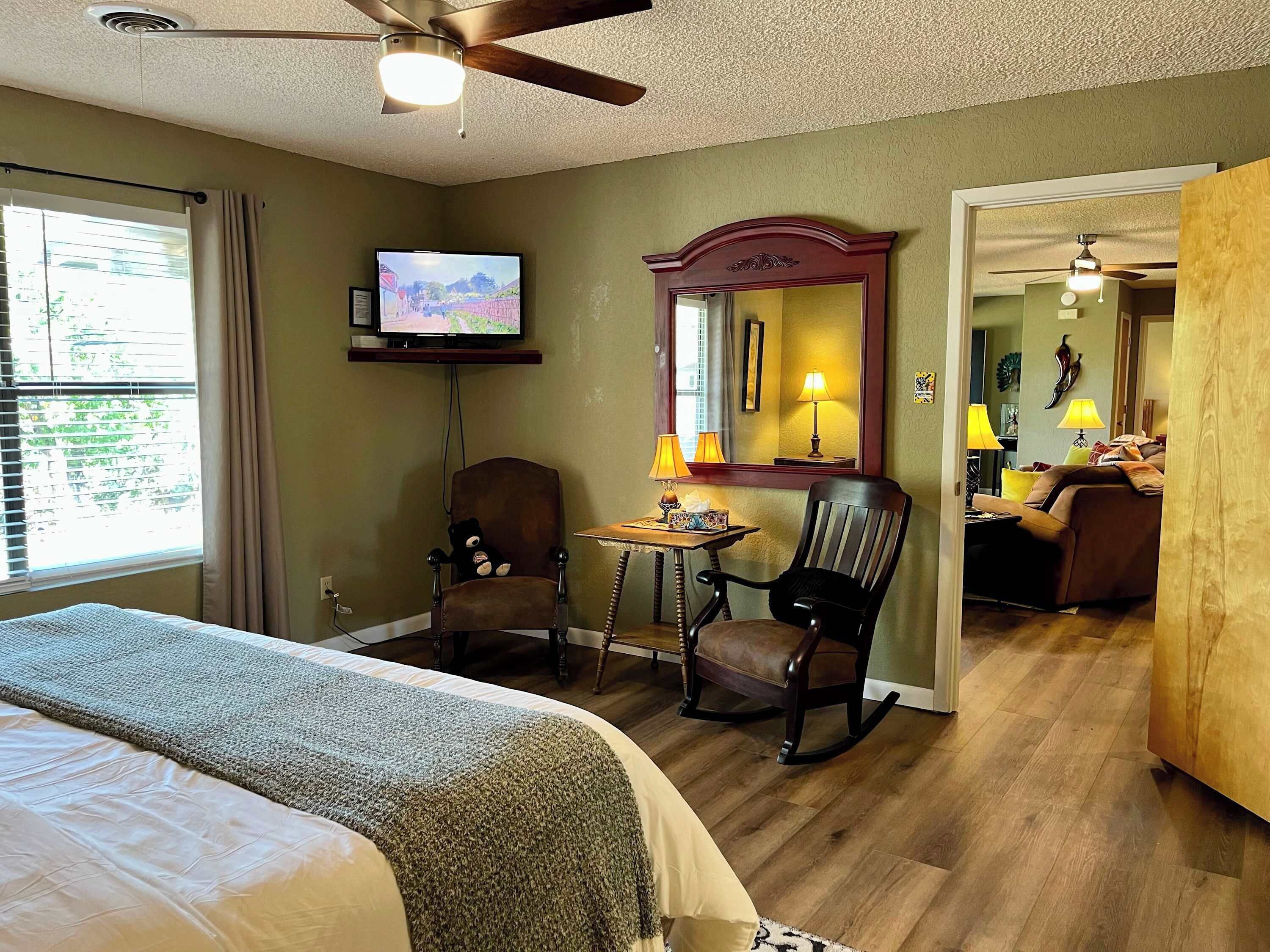 A hotel room with a bed, a desk with a chair, a television, a ceiling fan, and a large mirror.