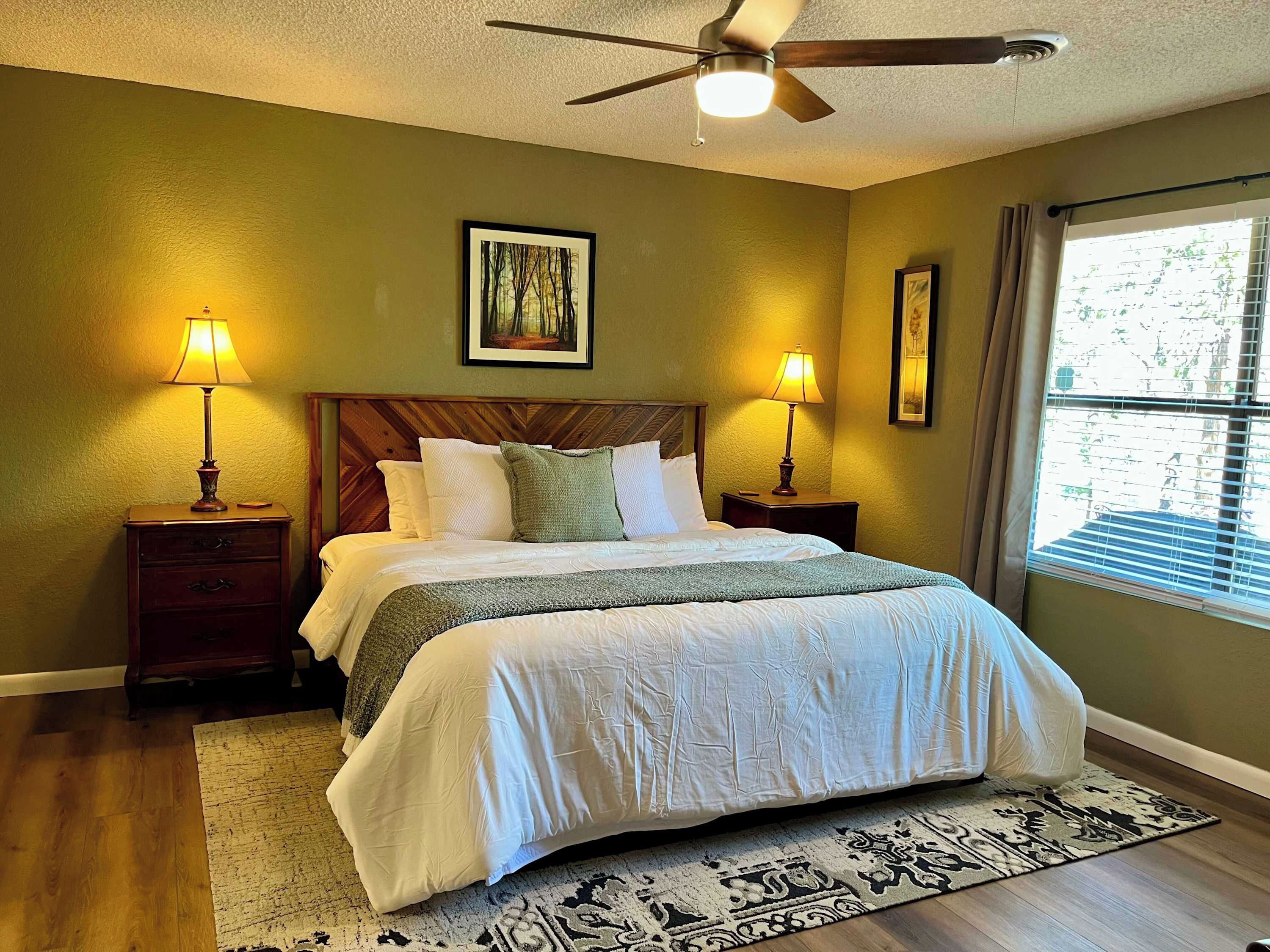A bedroom with a bed, two bedside tables with lamps, a ceiling fan, a window with curtains, and a framed picture on the wall.