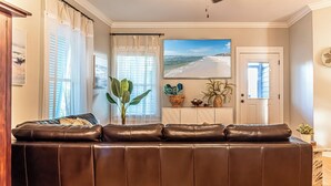 A living room with a brown leather sofa, a large window with sheer curtains, a potted plant, and a beach view painting on the wall. - 4005 Rookery 3 Sea Story (Gulf Shores)