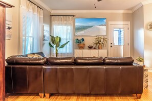 Ein Wohnzimmer mit einem braunen Ledersofa, einem großen Fenster mit transparenten Vorhängen, einer Topfpflanze und einem Gemälde mit Strandblick.