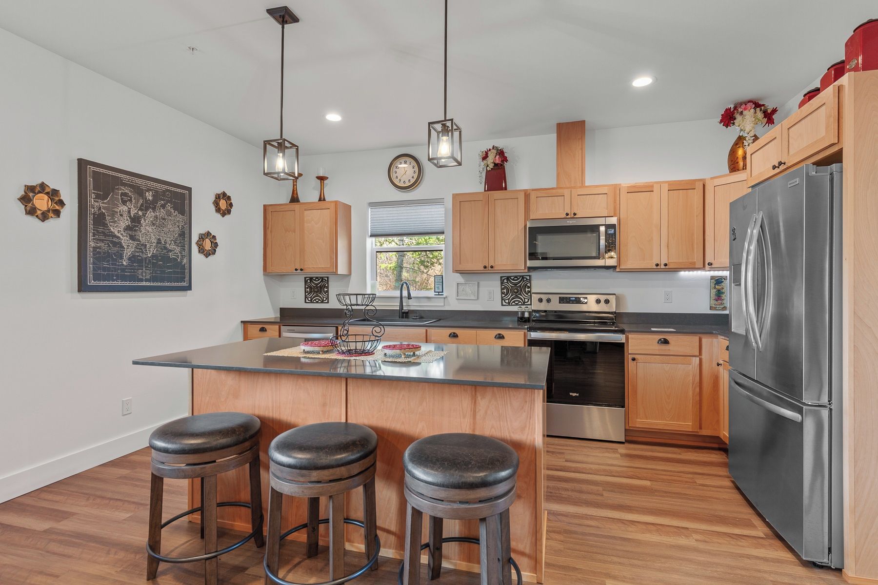 A modern kitchen with wooden cabinets, stainless steel appliances, a central island with stools, pendant lighting, and decorative wall art.