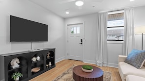 A modern living room with a flat-screen TV, a dark console table with decorative items, a round wooden coffee table with a green centerpiece, a white sofa with patterned cushions, and a window with curtains.