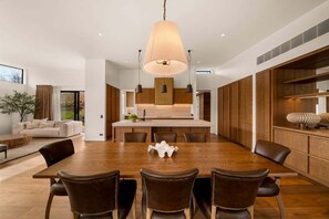 A modern dining area with a large wooden table, leather chairs, pendant lighting, and a view of the kitchen. - Villa Frogmore (Queenstown)