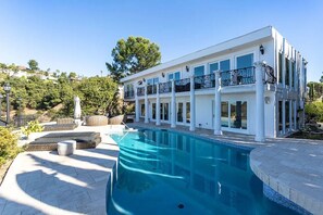Une maison moderne à deux étages, dotée d’une piscine, d’un espace salon extérieur et offrant une vue sur une colline.