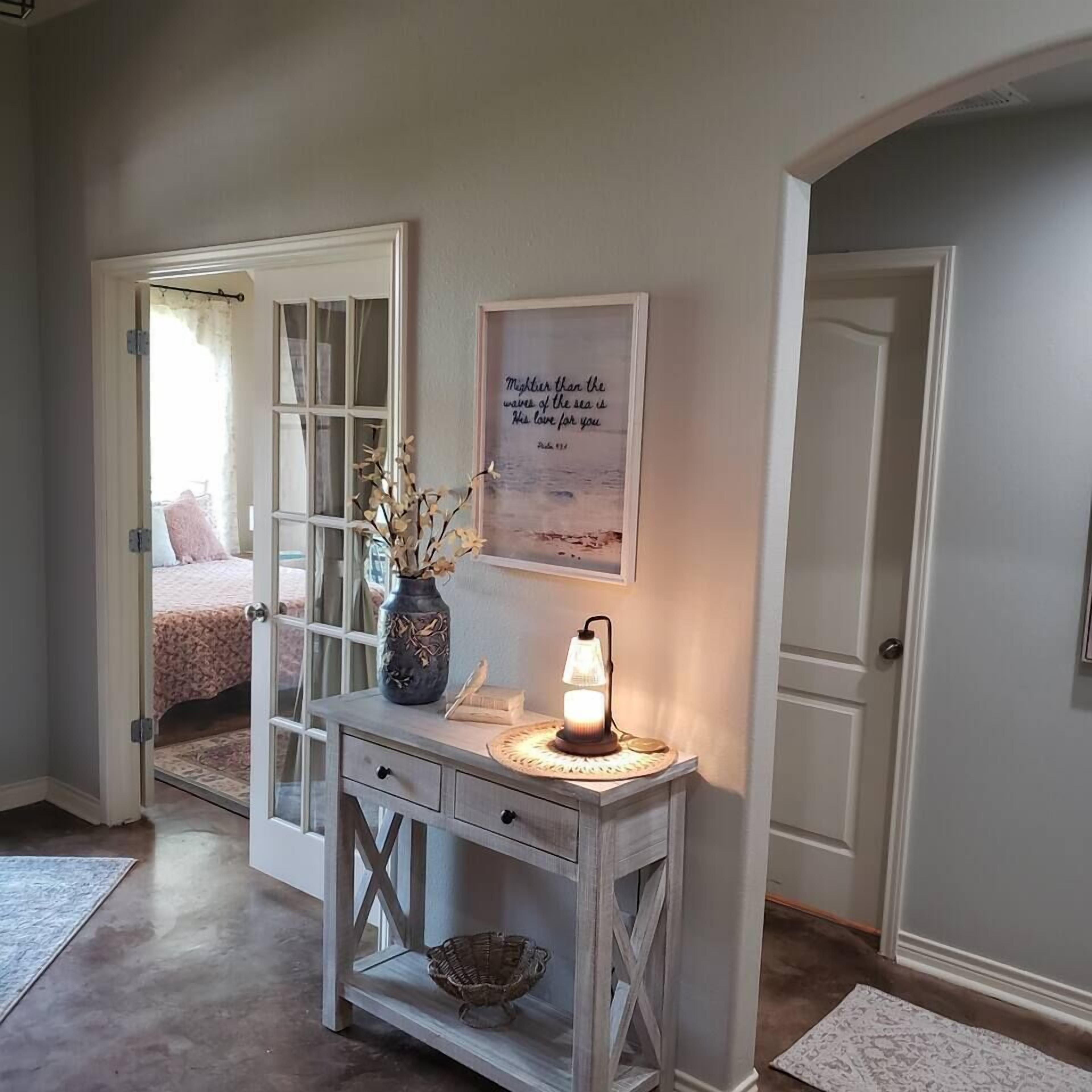 A room with a bed, a bedside table with a lamp, a framed quote on the wall, and a door leading to another room.