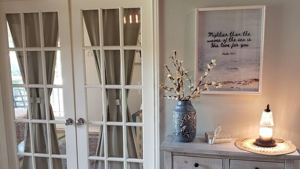 A room with a white door with glass panels, a small table with a vase and a lamp, and a wall-mounted picture with a quote. - Spacious Bixby Home w/ Fire Pit & Sleeps 12+ (Bixby)