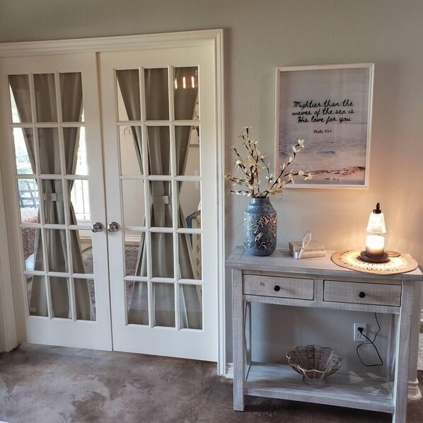 Ein Zimmer mit einer weißen Tür mit Glasfeldern, einem kleinen Tisch mit einer Vase und einer Lampe sowie einem an der Wand hängenden Bild mit einer Zitat.