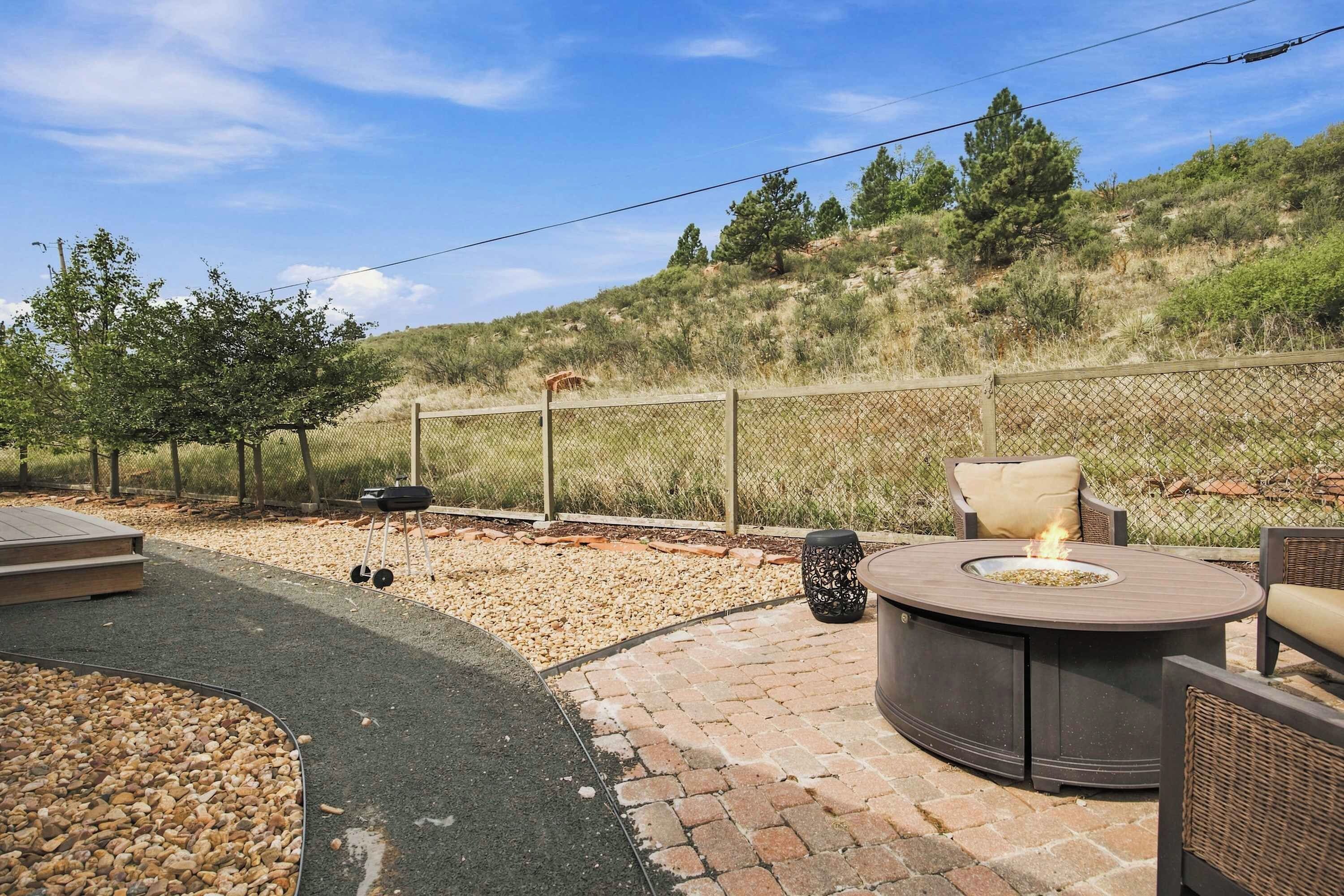 An outdoor patio with a round table and chairs, a grill, and a fenced area with a hilly landscape in the background.