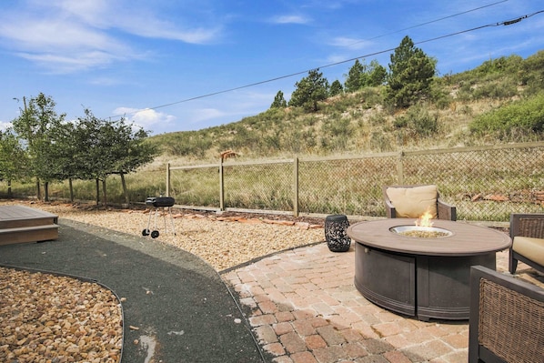 An outdoor patio with a round table and chairs, a grill, and a fenced area with a hilly landscape in the background. - Dixon Cove Lookout Near Horsetooth Res (Fort Collins)