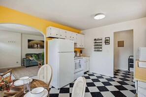 Eine kompakte Küche mit weißen Schränken, einem Kühlschrank und einem Herd. Es gibt eine Essecke mit einem Tisch für zwei Personen, und ein Schachbrettmuster auf dem Boden.