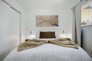 Un lit bien fait, avec une couette blanche et un arrangement décoratif de couvertures, un tableau de fleurs accroché au mur et deux lampes de chevet.