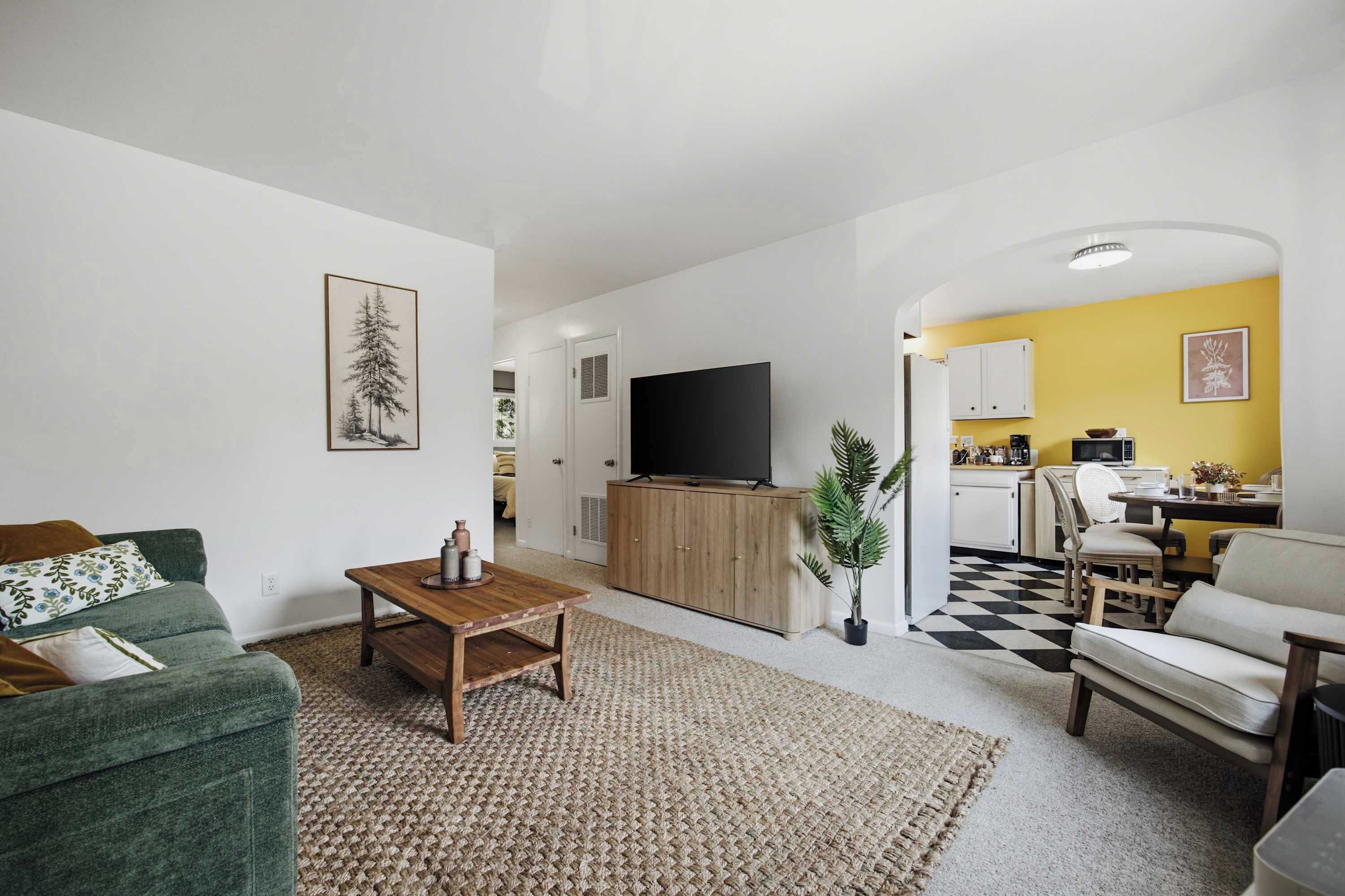 Ein Wohnzimmer mit Sofa, Couchtisch und Fernseher. Es gibt eine Küchenzeile mit Esstisch und Stühlen.