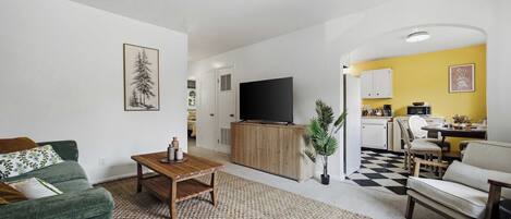 Una sala de estar con sofá, mesa de centro y televisor. Cuenta con una zona de cocina con mesa y sillas.
