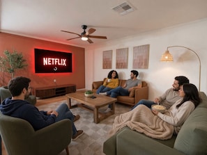Ein Wohnzimmer mit einem Flachbildfernseher, der 'NETFLIX' anzeigt, einem Deckenventilator und einem Couchtisch mit einer Schüssel Popcorn.