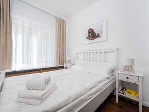 A neatly made bed with white linens and a rolled towel, a bedside table with a lamp, a small bedside table with a book and a remote control, and a window with sheer curtains. - Manufaktura Cosy Apartment (Łódź)